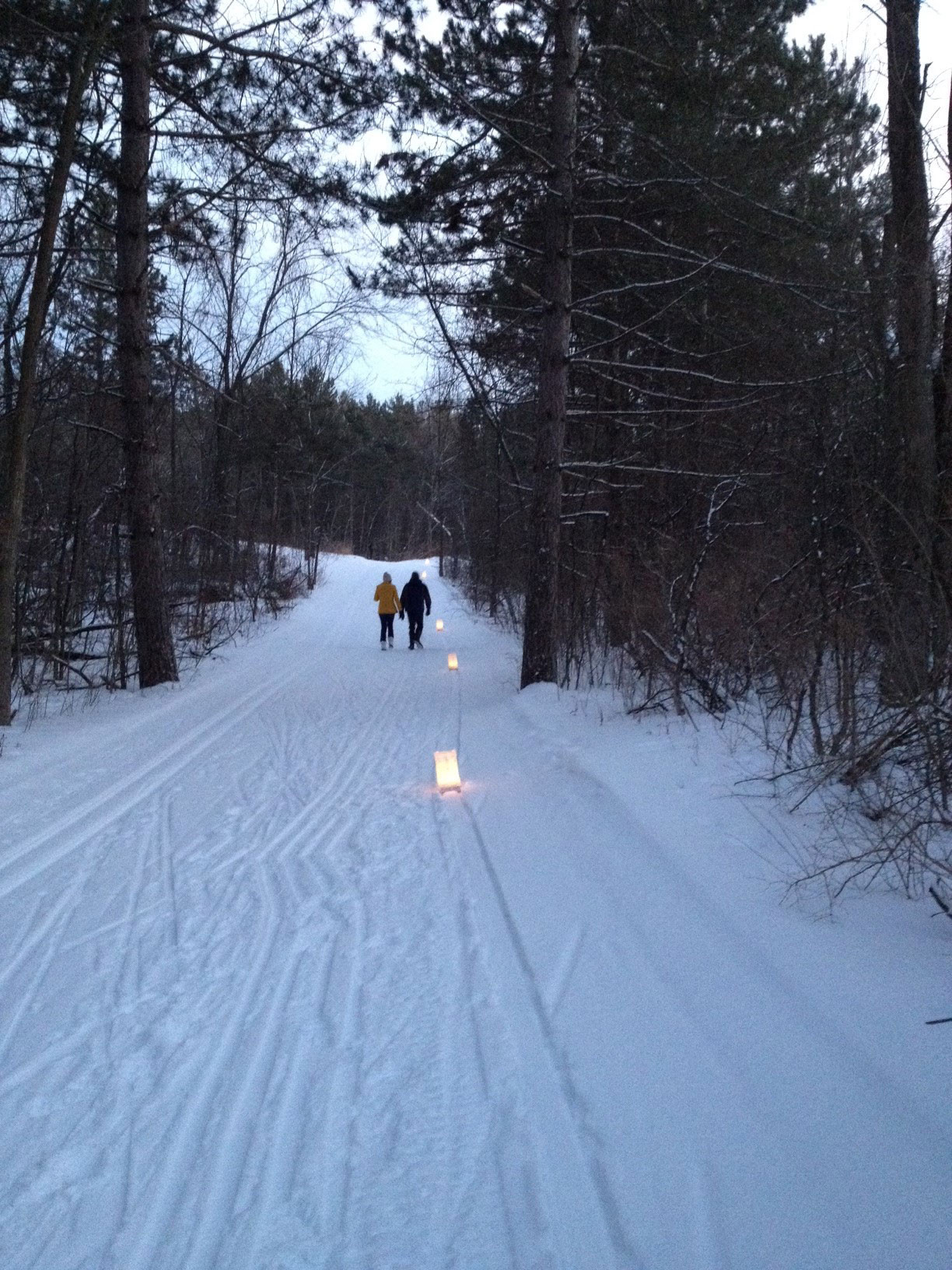 Two figures walk on a snowy trail at twilight lit by candle luminaries.