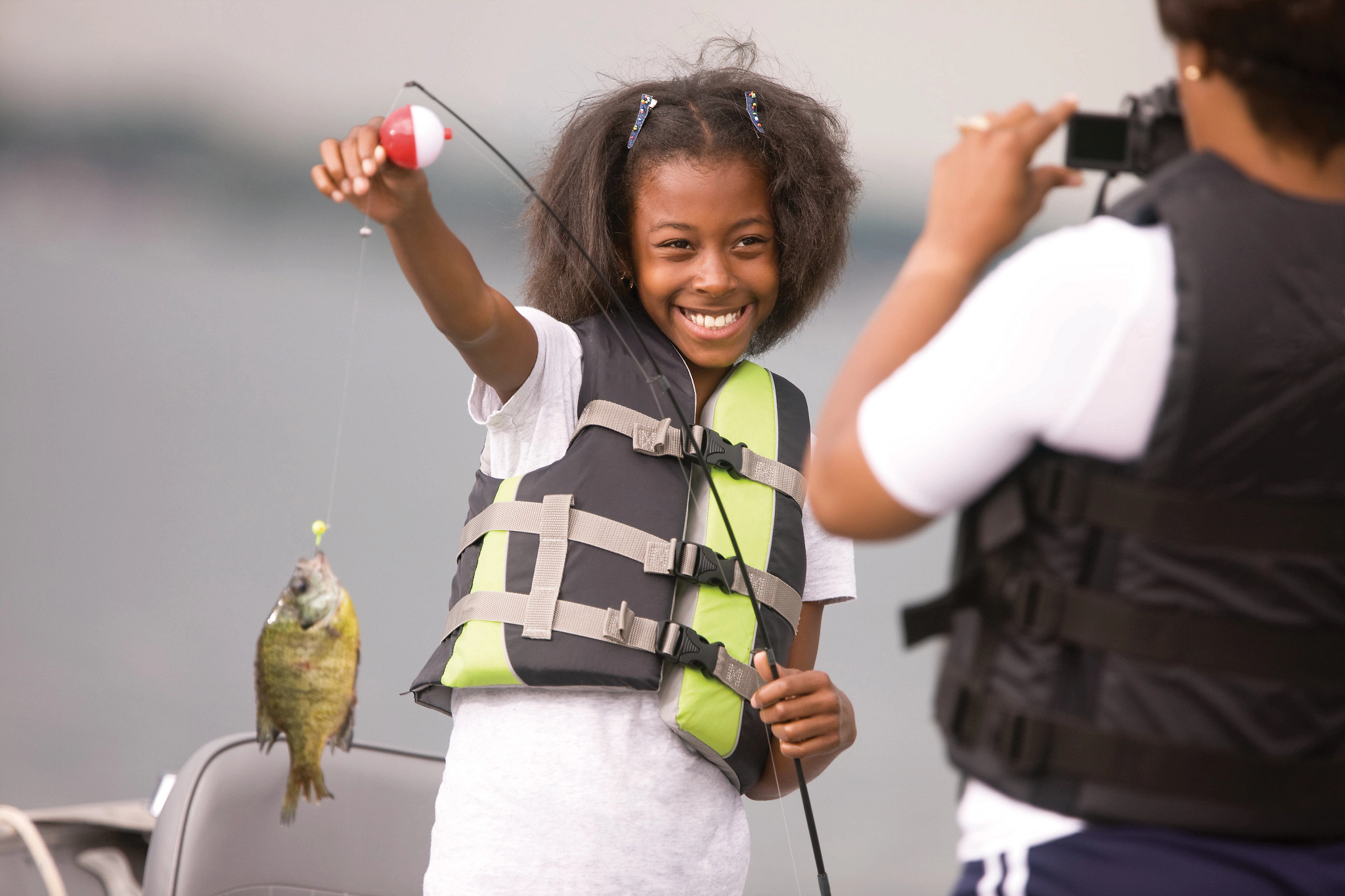 A young black female angler holds up a fish while an older woman takes a photo of her.