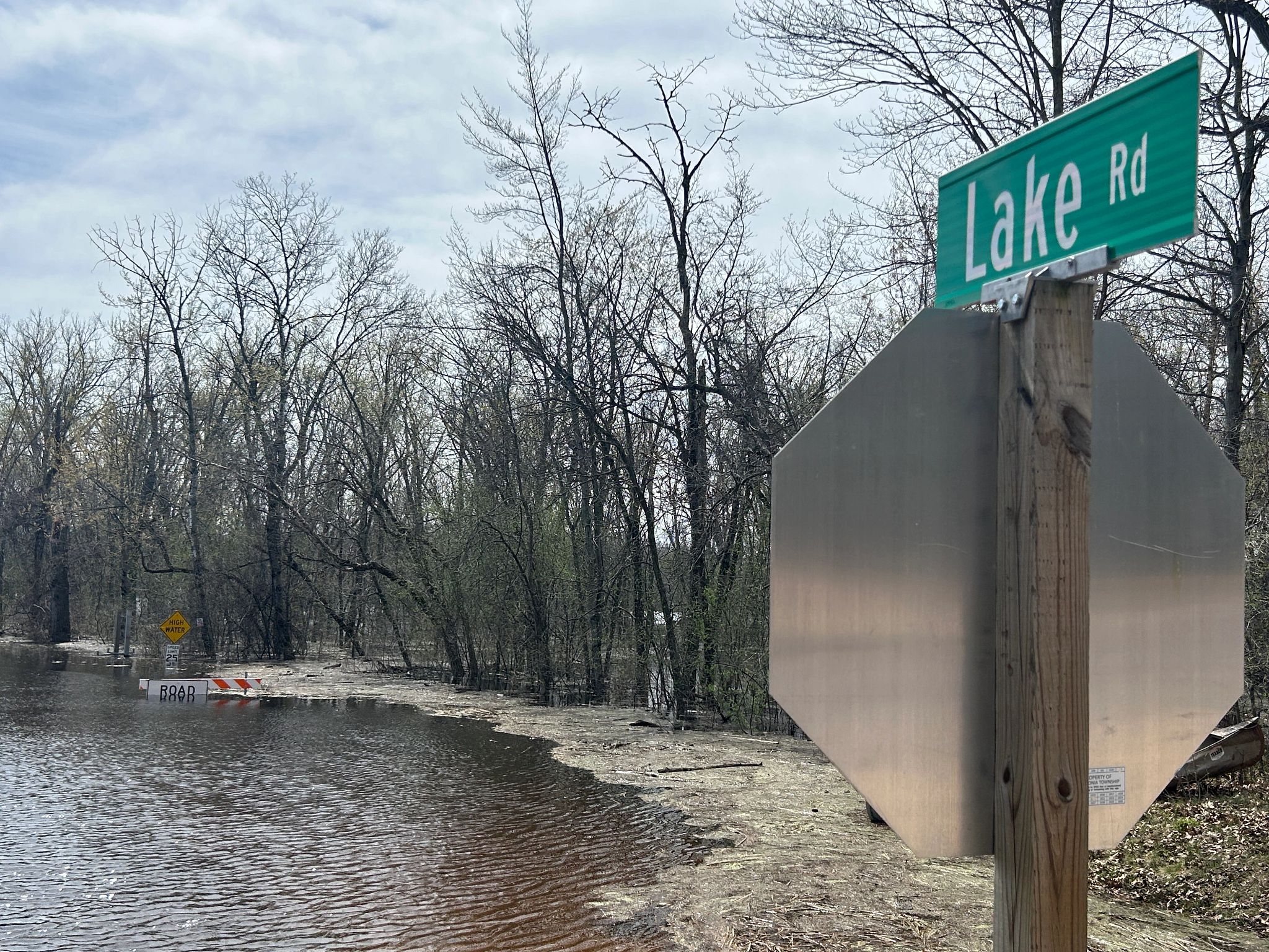 Flood waters over a road flanked by trees. A stop sign with a street sign that says Lake Road is in the foreground.
