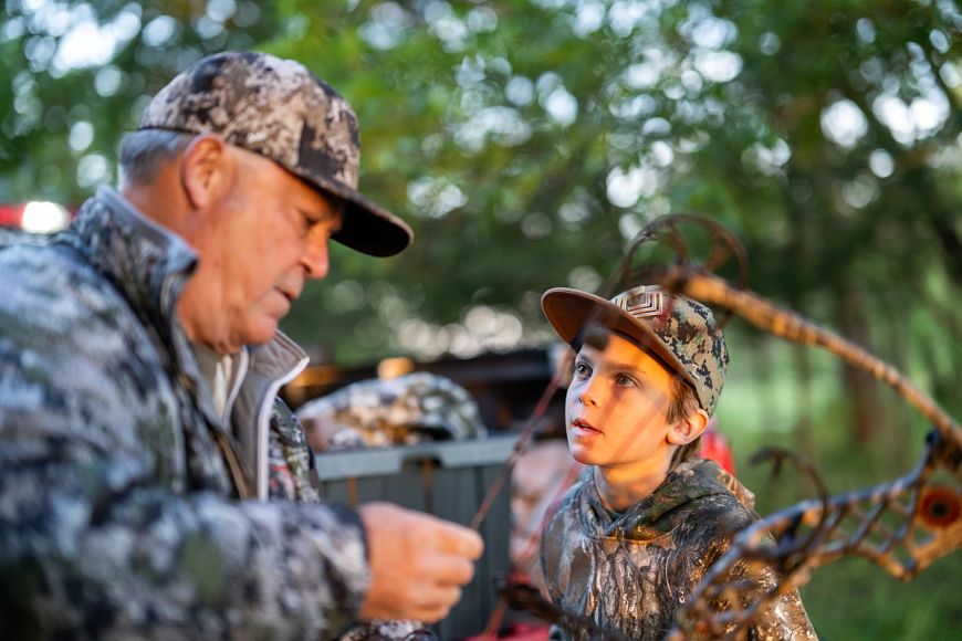 A man in camo holds a bow as a young boy in camo looks on.