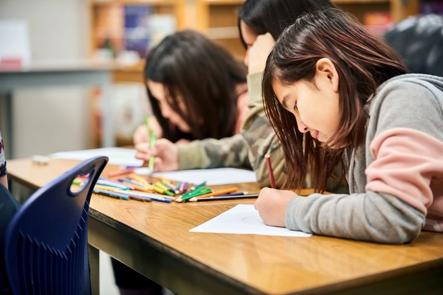 Three students hunch over at a desk drawing pictures with colored pencils.
