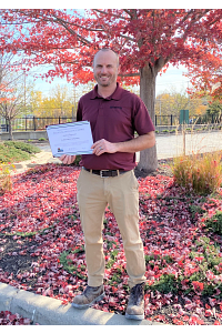 A Kaukauna Utilities representative stands with their award in front of a tree with red leaves.