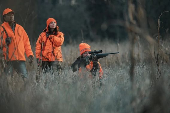 A woman aims her rifle as two people stand behind here. All are wearing Blaze Orange.