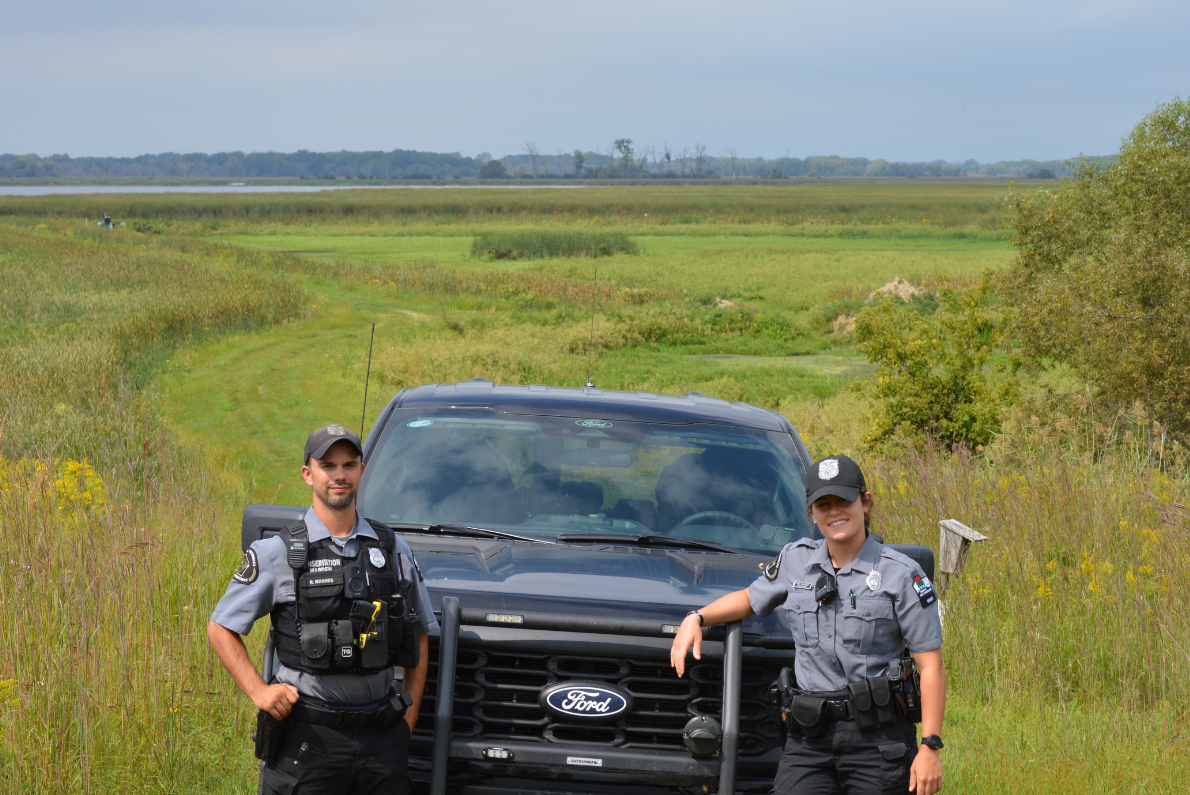 Two wardens pose, flanking a warden truck that is parked in front of an early-spring, green meadow. 