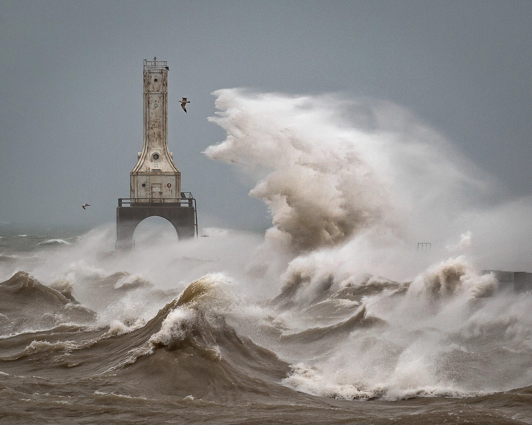 A monster wave crashes over the breakwater towards the Port Washington Lighthouse. The crest of the wave looks a bit like Godzilla, mouth agape and ready to swallow the seagull flying frantically away and maybe the lighthouse too. Below the great wave, Lake Michigan churns chaotically with frothy taupe and white waves, against a misty leaden grey sky.