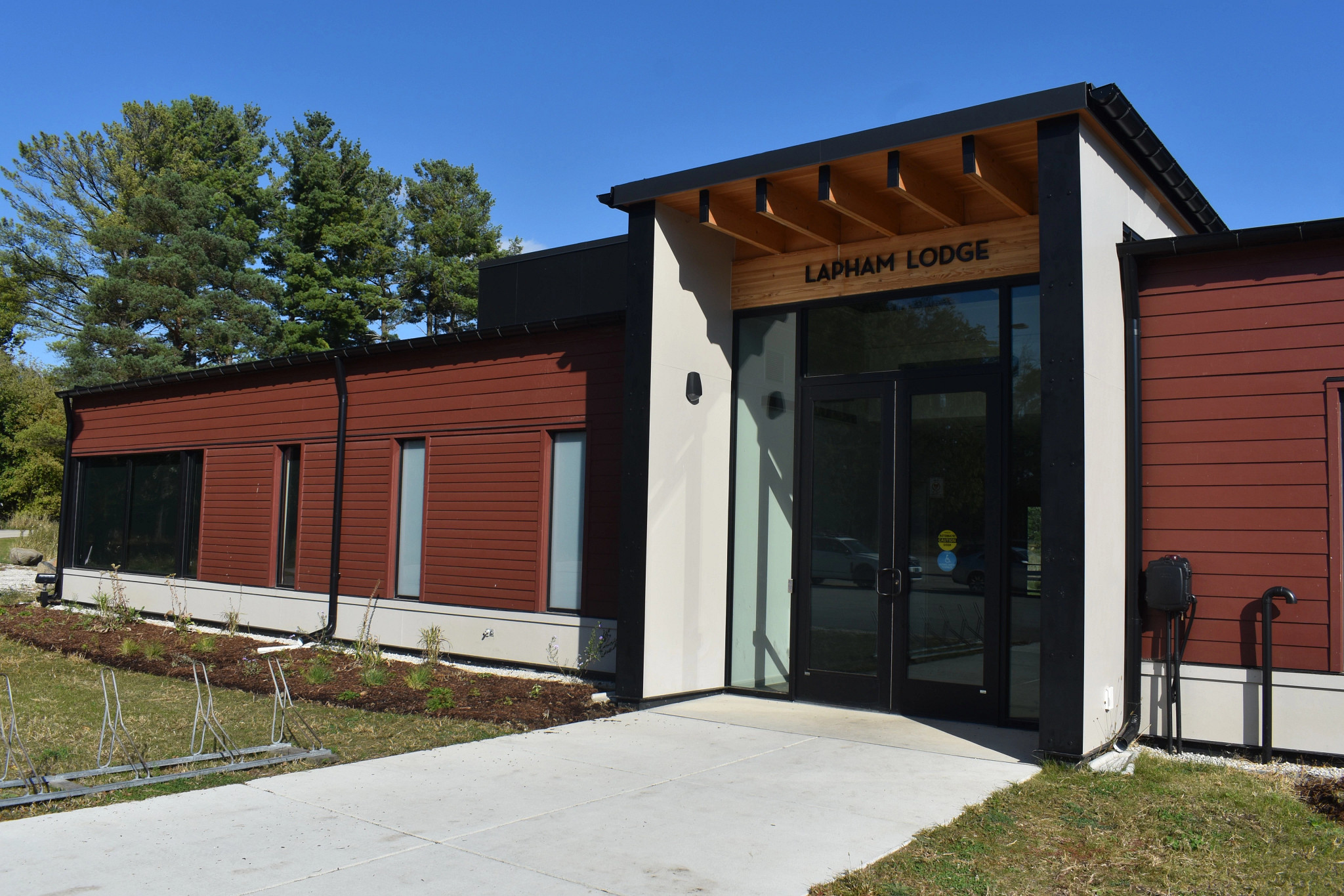 This image shows the exterior of the Lapham Lodge, a modern building with dark red siding and large windows, surrounded by grass and trees under a clear blue sky.
