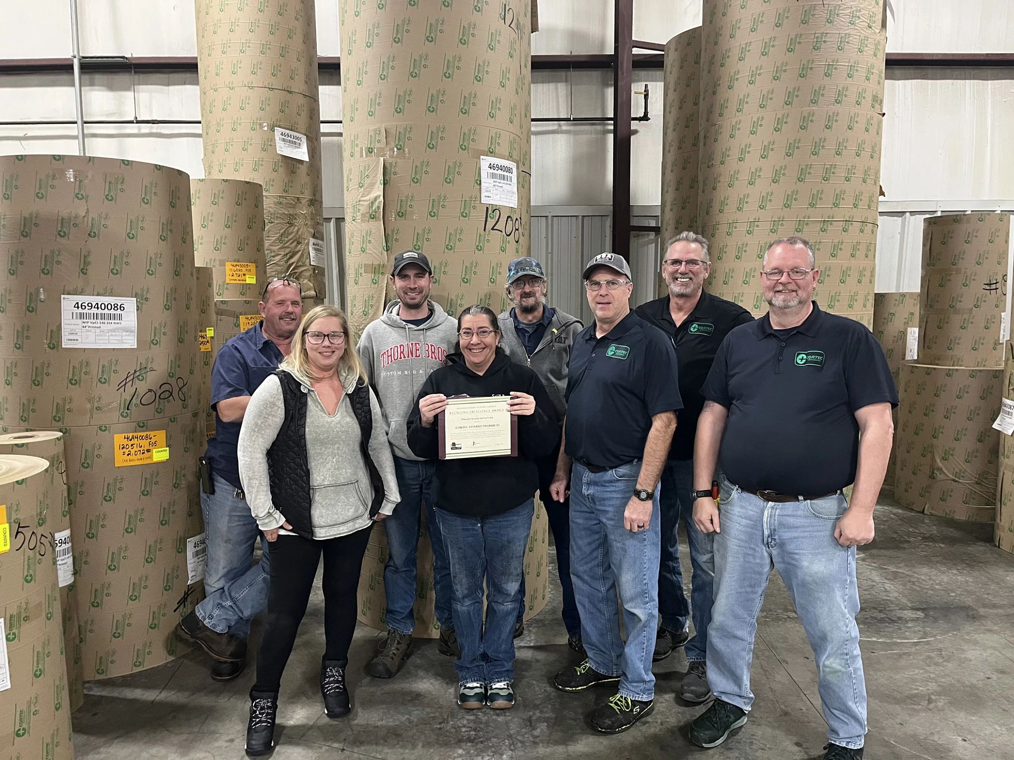 The team at Cortec Coated Products gathers together in front of giant rolls of coated paper. The team member in the center holds the business's 2023 Wisconsin Recycling Excellence Award certificate from the DNR.