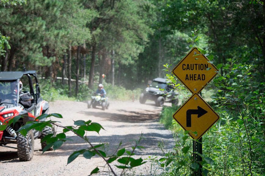 A group of UTVs and ATVs ride on a dirt trail through the woods. A sign reading "Caution Ahead" is seen in the foreground on the side of the trail.