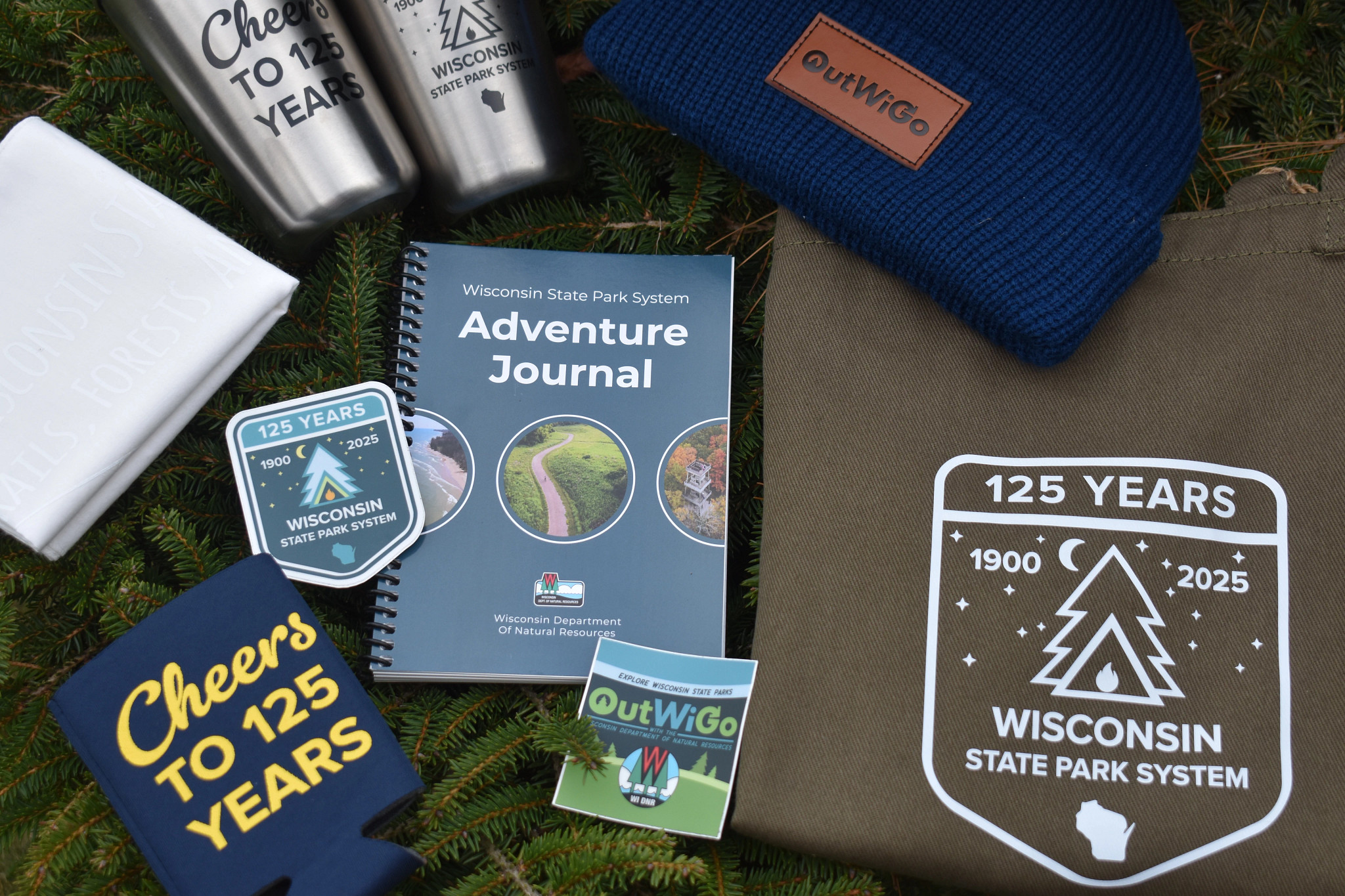 DNR 125th anniversary Parks merchandise laying on a table, including an Adventure Journal, hat, coozie and T-shirt
