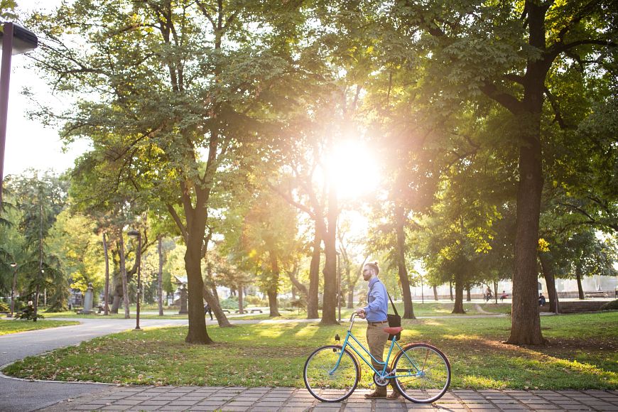 A man wearing business casual clothing pushes a bike along a cobblestone path. The sun shines through the green trees in the background, illuminating the green space next to his path.