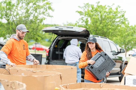 Two people, both in orange shirts, place old electronics into cardboard bins for sorting prior to recycling.