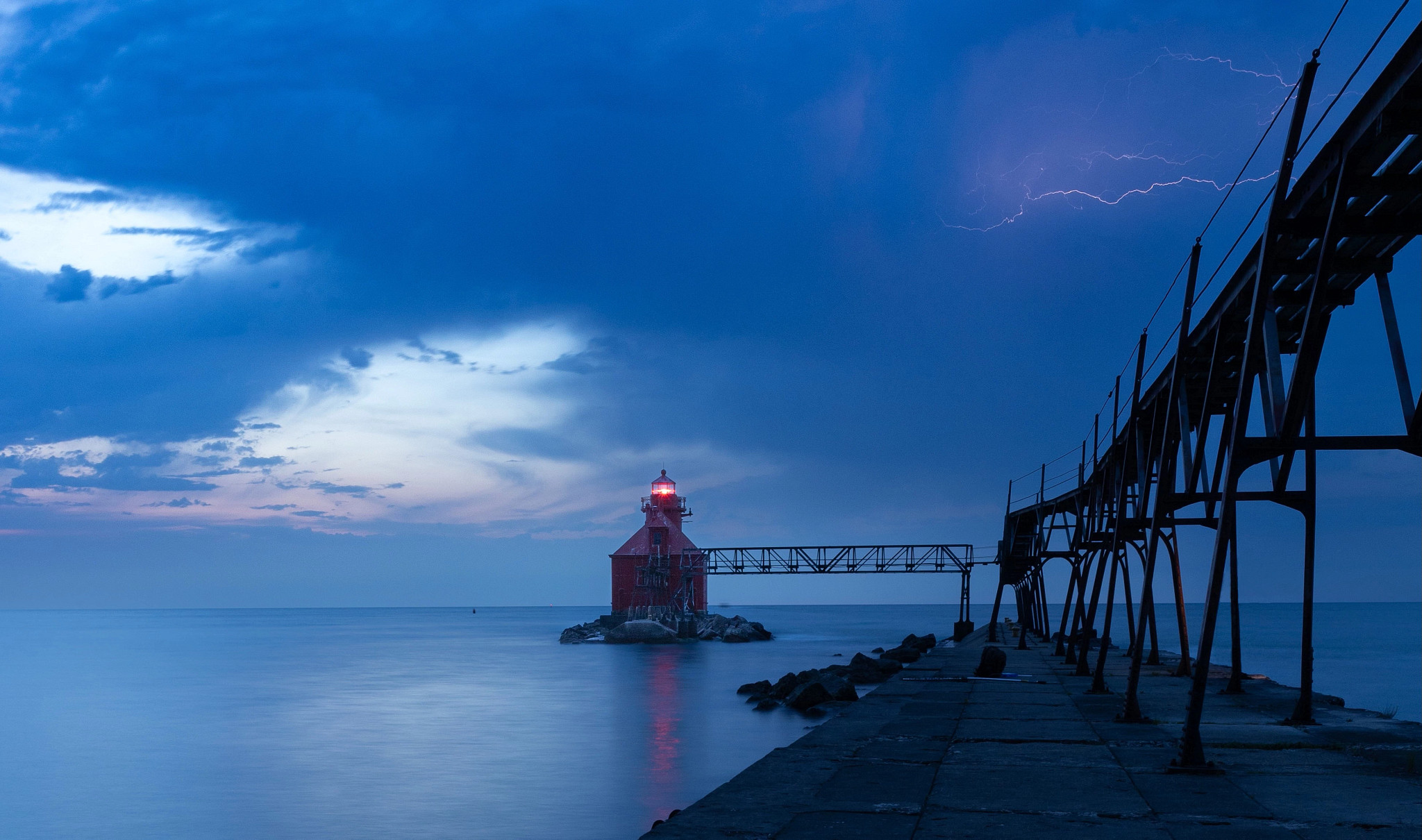 Lightning makes jagged pink streaks across the southern sky, seeming to come out of the catwalk leading to the Sturgeon Bay Ship Canal Pierhead Lighthouse. The station light glows red against a moody dark blue sky at the end of the catwalk. The light casts a red reflection on the eerily calm water, while a clearing patch in the northeastern sky to the left of the lighthouse shows a hint of dawn’s soft pink light.
