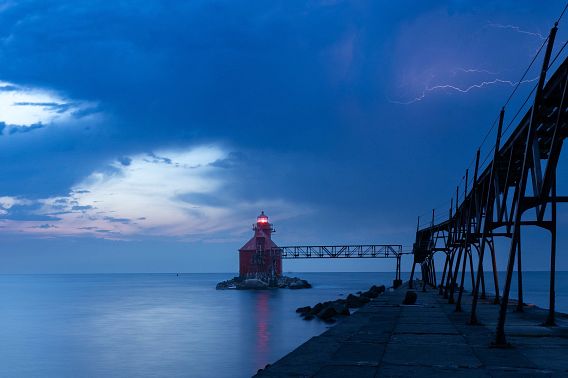 Lightning makes jagged pink streaks across the southern sky, seeming to come out of the catwalk leading to the Sturgeon Bay Ship Canal Pierhead Lighthouse. The station light glows red against a moody dark blue sky at the end of the catwalk. The light casts a red reflection on the eerily calm water, while a clearing patch in the northeastern sky to the left of the lighthouse shows a hint of dawn’s soft pink light.