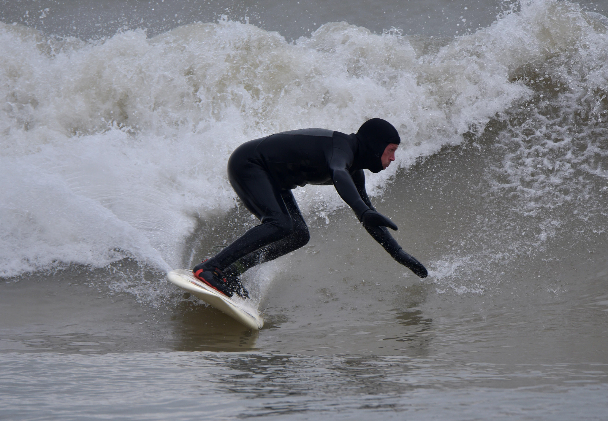 A surfer in a hooded wetsuit crouches with perfect balance, edging his surfboard, as he glides down the face of a rolling wave on Lake Michigan. His mittened left hand nearly touches the water in front of him as he gazes with keen focus on the arc of his path down the wave. The crest of the wave is a frothy grey and white wall splashing high above his head.
