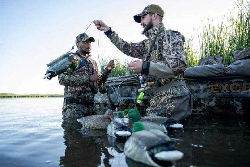 two waterfowl hunters in water near a body holding decoys