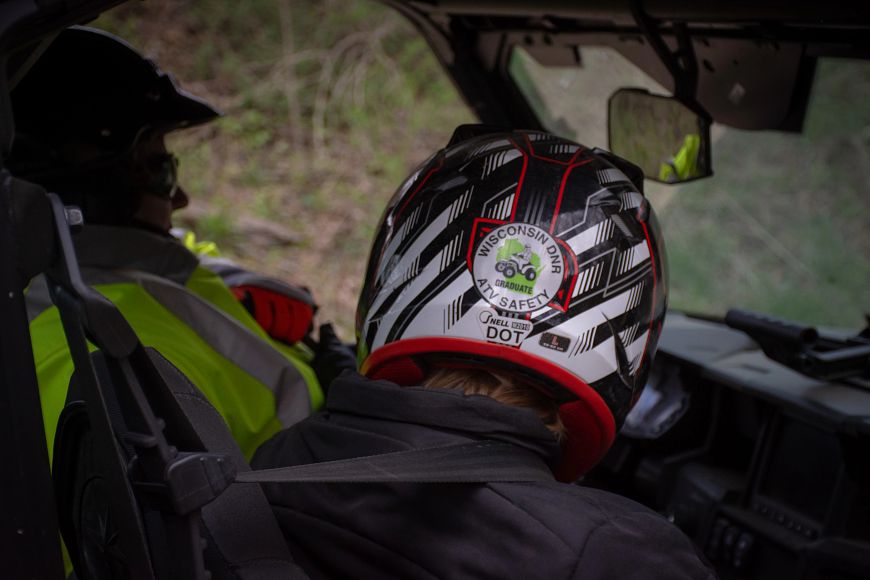 The back of a helmet on the passenger of a UTV is seen, with a sticker that reads "Wisconsin DNR ATV Safety." The driver of the UTV is seen over the left shoulder of the passenger. 