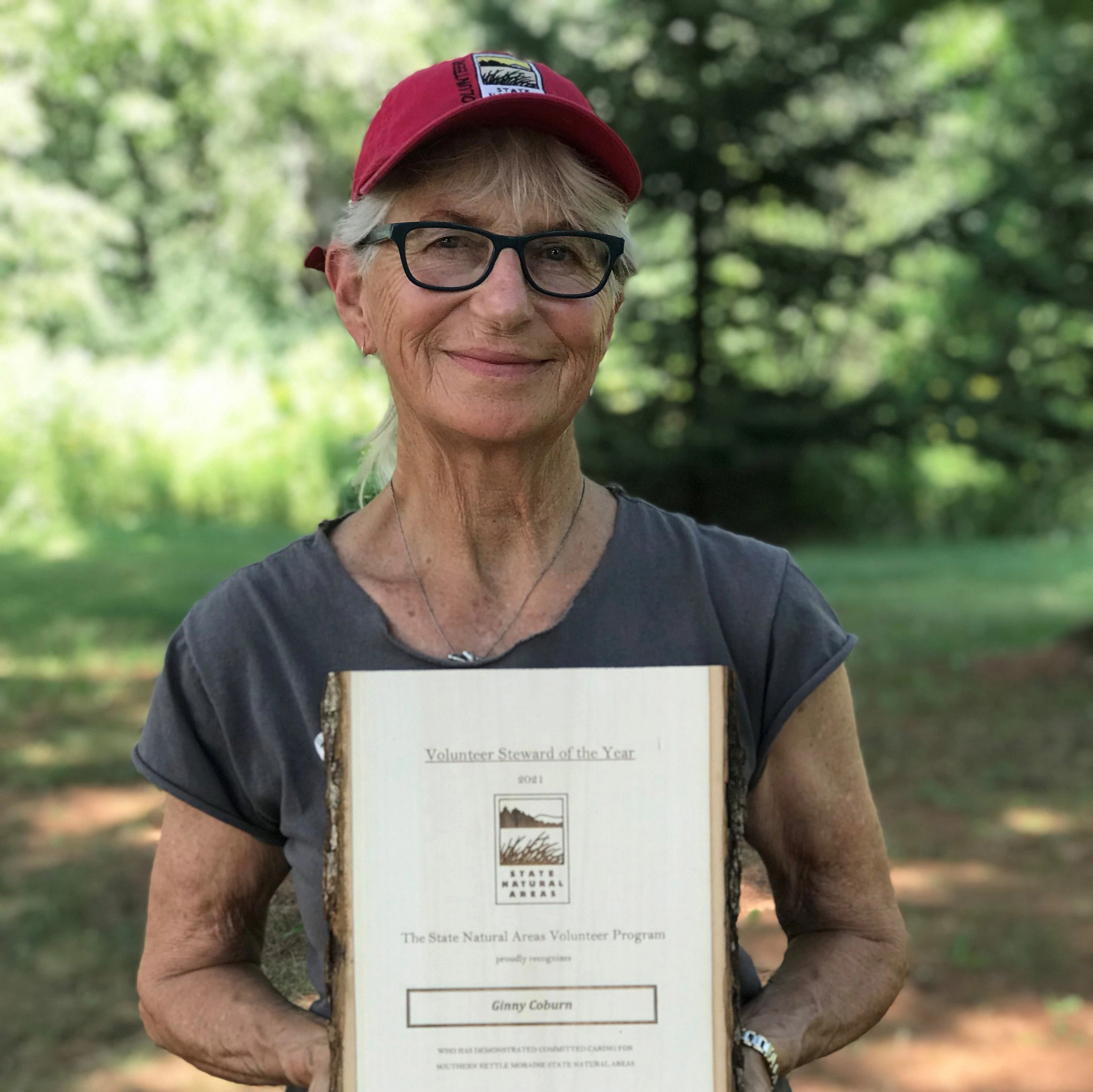 Ginny Coburn, with light skin tone, gray hair, a red baseball cap and glasses, smiles holding up her Volunteer Steward of the Year award. 