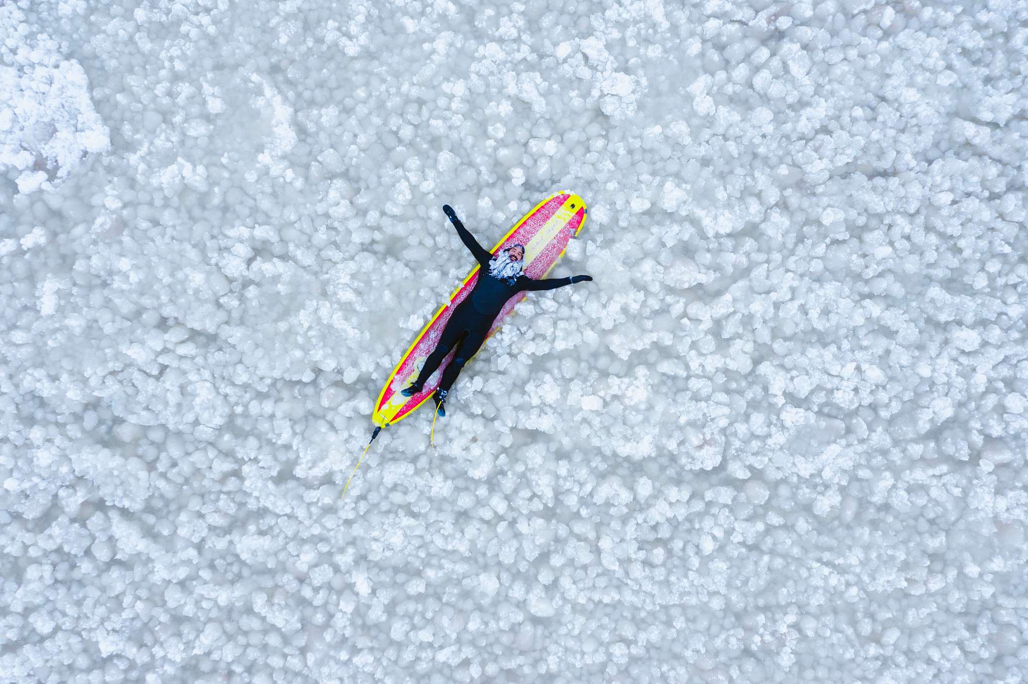 A man in a black wetsuit with an icicle beard laying on a yellow and red striped surfboard in a frozen lake.