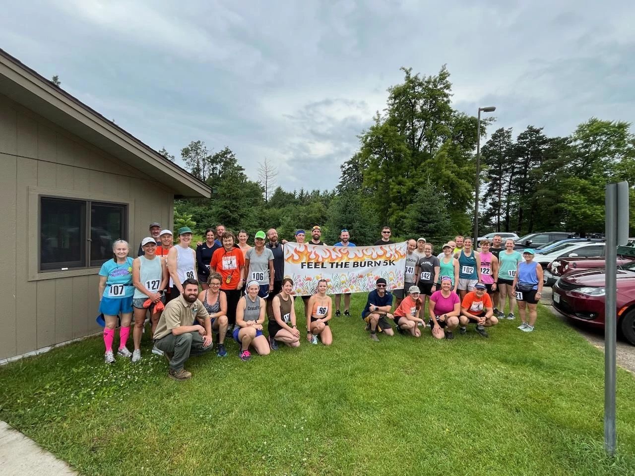 Group of people posing outdoors for the “Feel the Burn 5k” event holding a banner with trees and cars in the background.