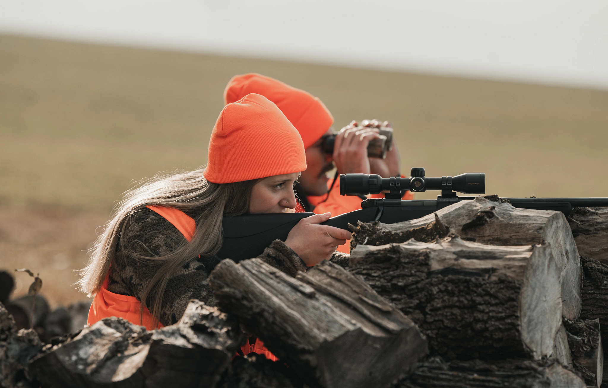 Two hunters in orange hats are using a gun and binoculars to scan the area, positioned behind a log in a field.