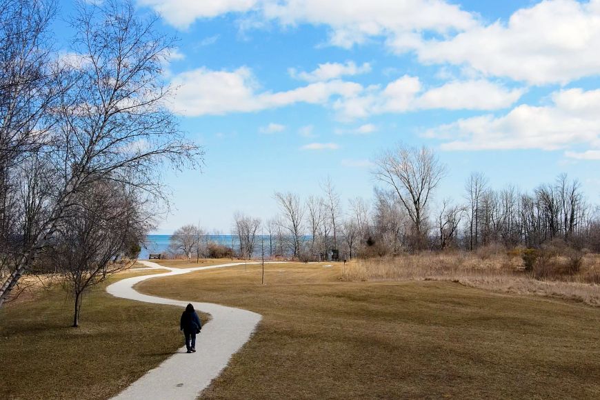 An aerial view of an individual walking along a paved path winding through an open field of brown grass, with a large lake and clear blue sky visible in the distance.