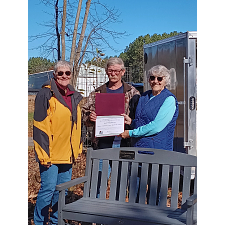 Three participants stand outside holding their award next to a bench created from recycled plastic