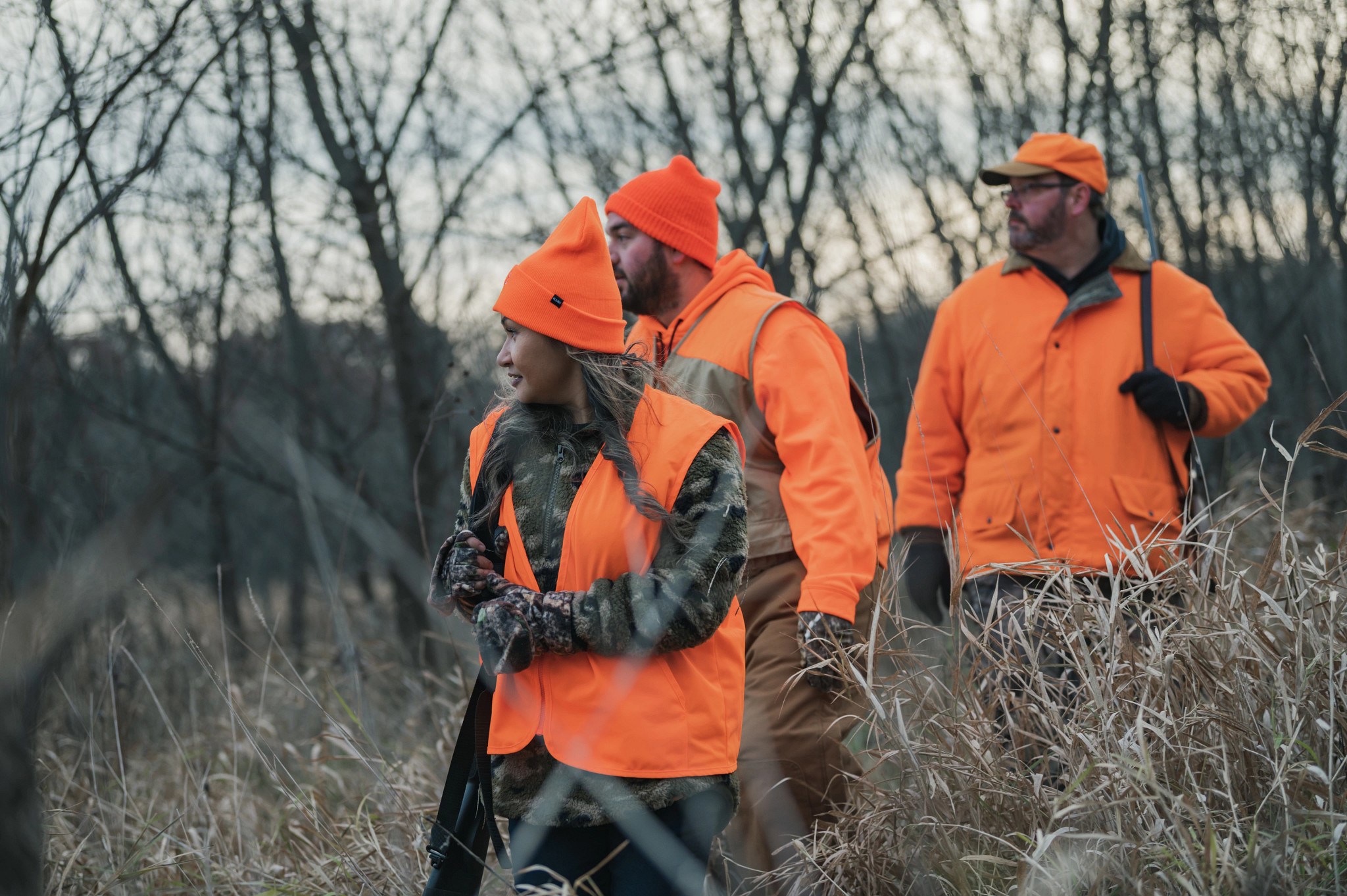 Three deer hunters walk through a field of tall grass safely shouldering their firearms and wearing blaze orange.