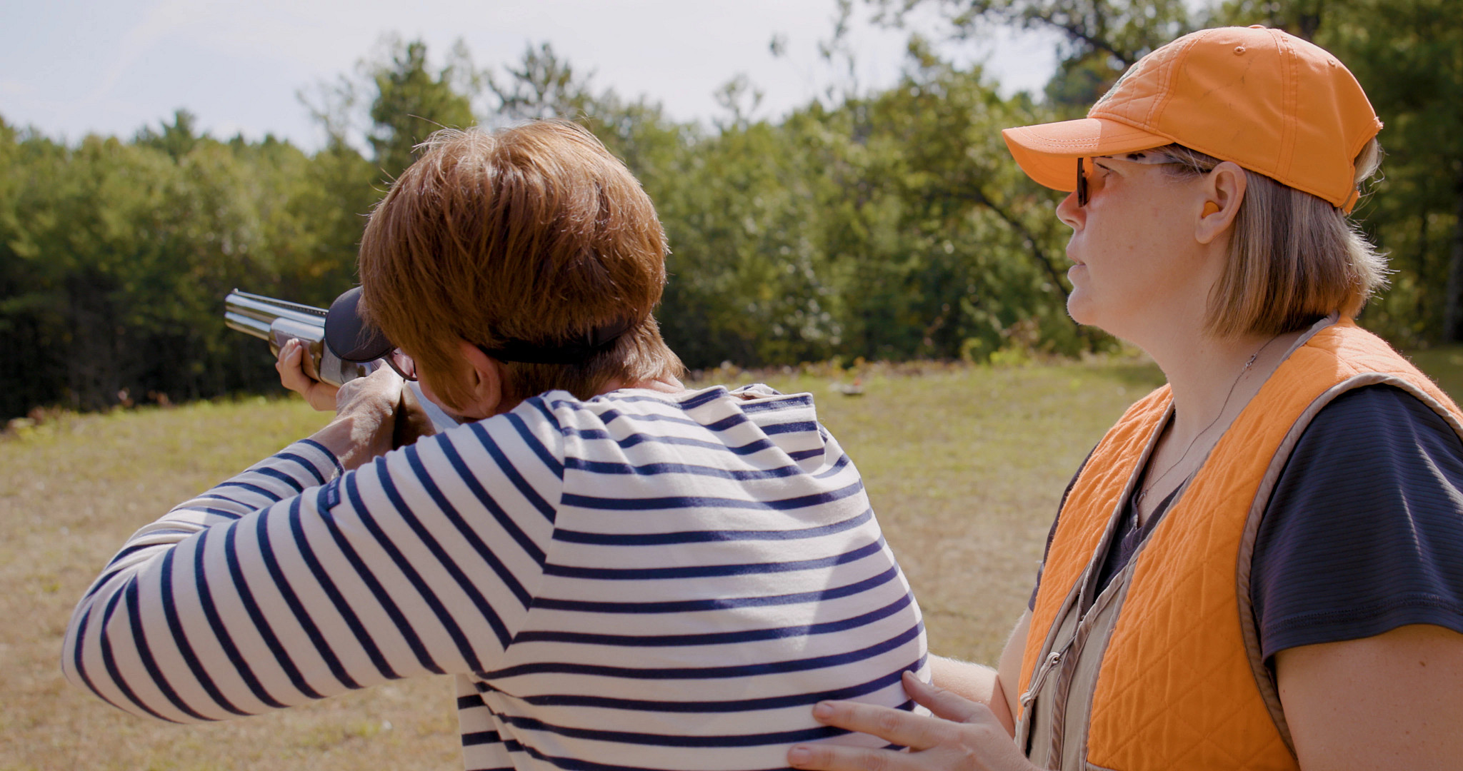 hunter ed instructor wearing blaze orange helps a woman aim a gun at a shooting range