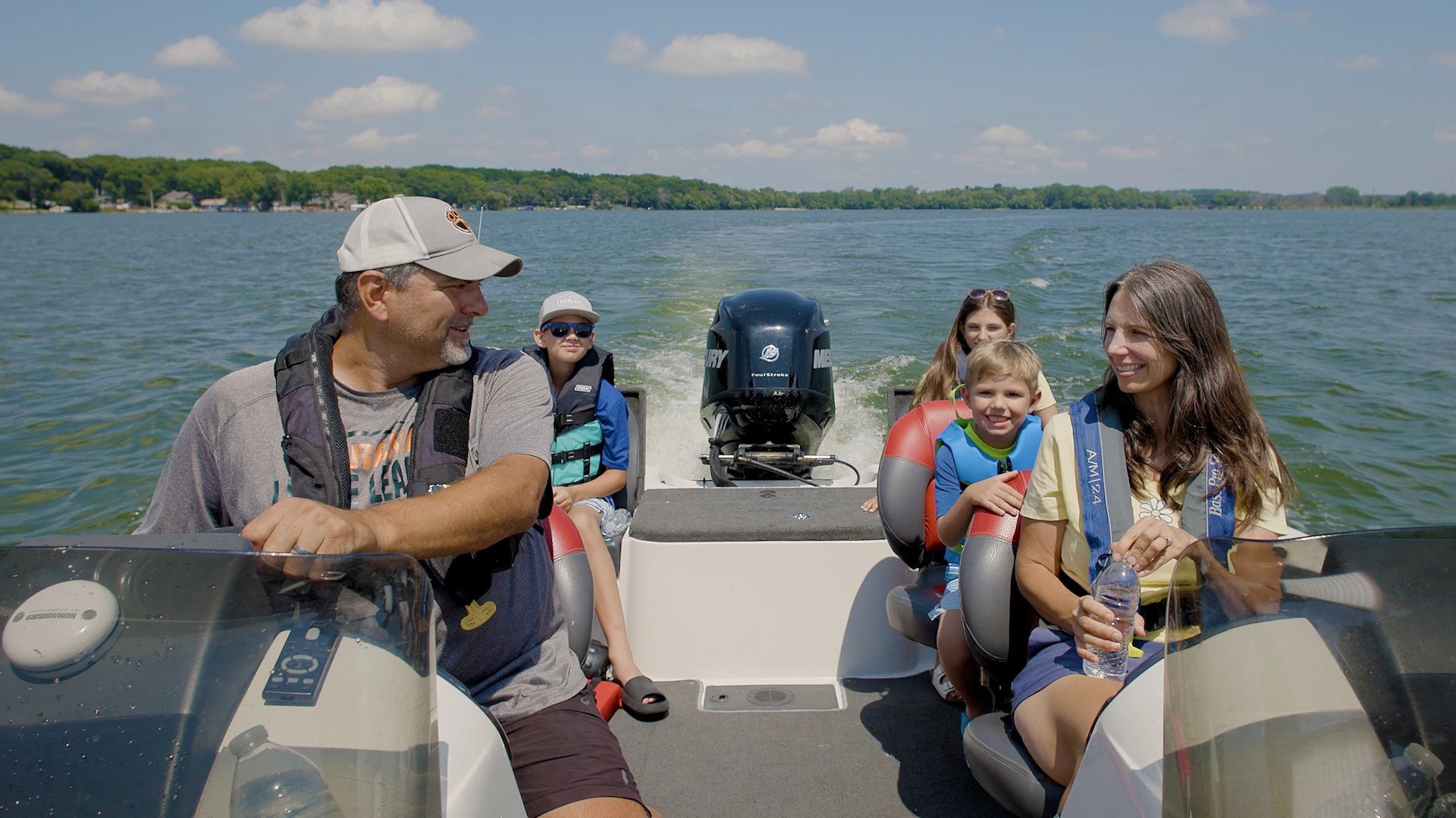 A family of five, all wearing life jackets, enjoying a boat ride on a sunny day. 