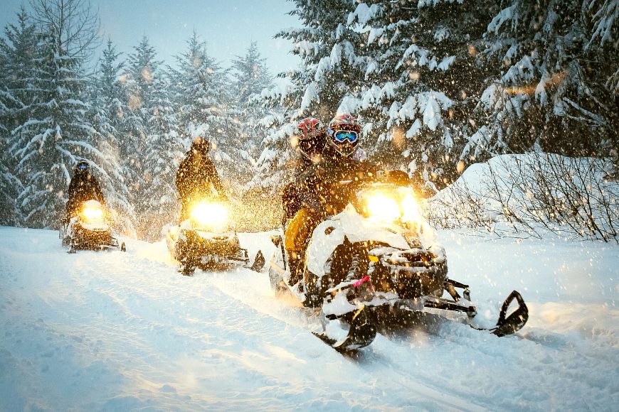 Three snowmobiles with their lights on are ridden through the falling snow on a wooded trail. 