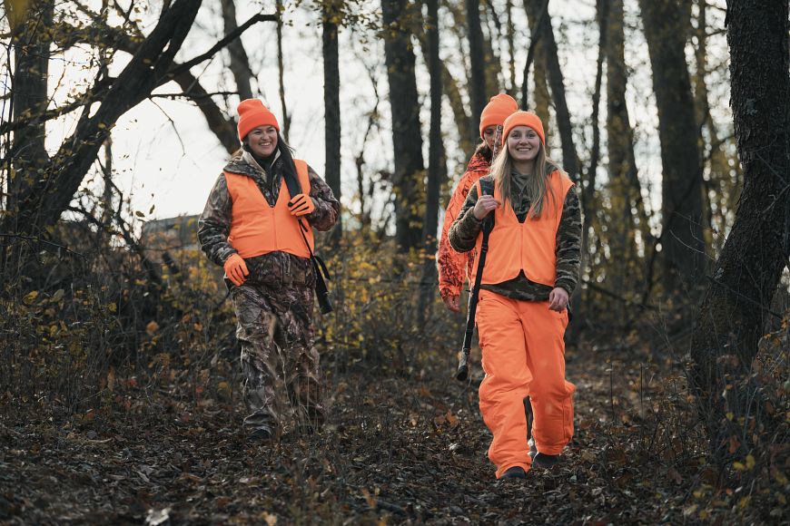 Three female hunters wearing camouflage and blaze orange, walking in the woods.