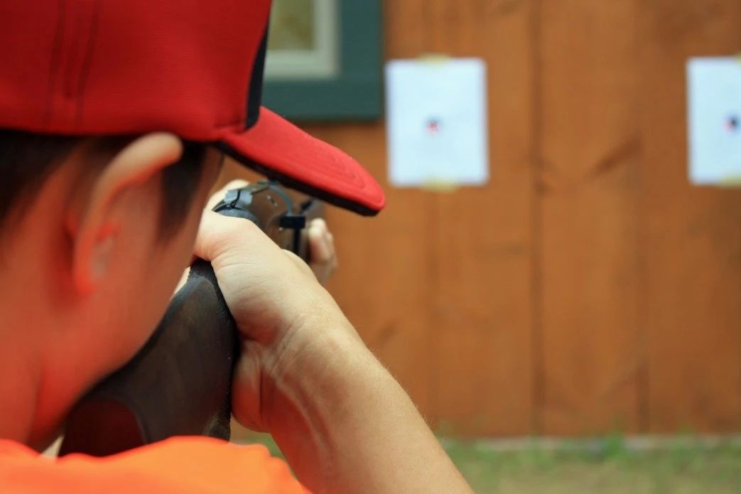 A young boy wearing a hat and blaze orange shirt, viewed from behind his shoulder, aims his rifle at a target.