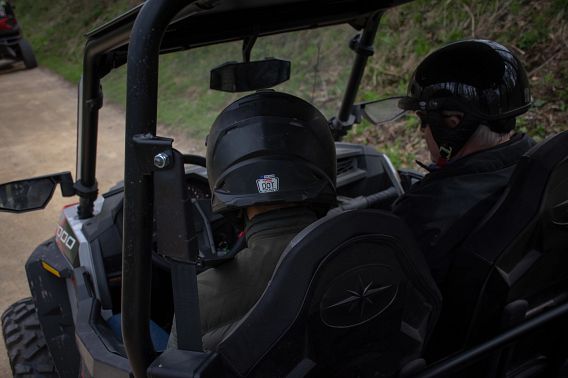 Two men, both wearing helmets and dark jackets, ride in a UTV on a wooded trail.