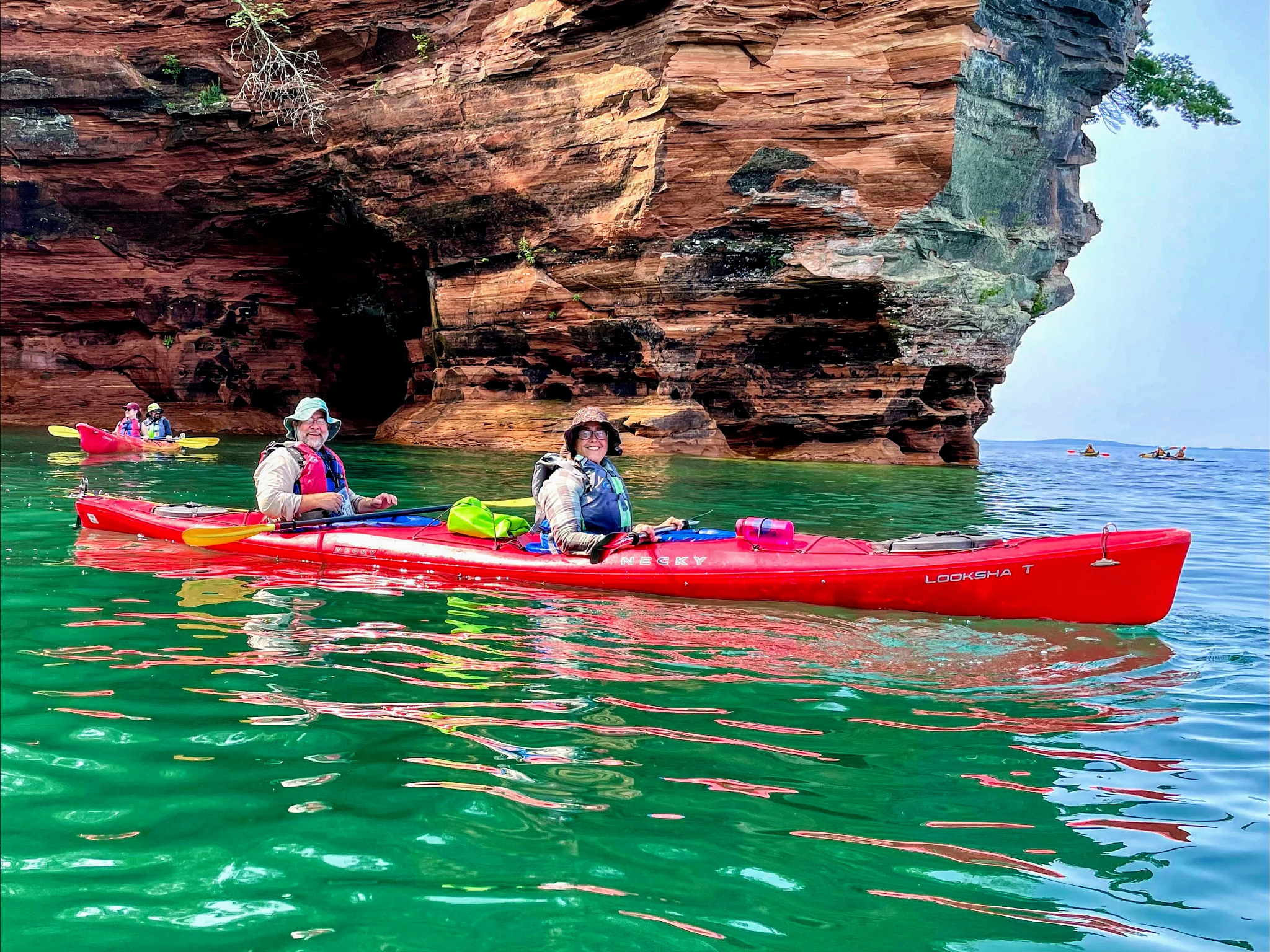 Two smiling people in a tandem sea kayak pause in calm water in front of a tall sandstone sea cave. Behind them, another duo in a tandem sea kayak pause near the sea cave. Another set of kayakers are paddling around the headland.