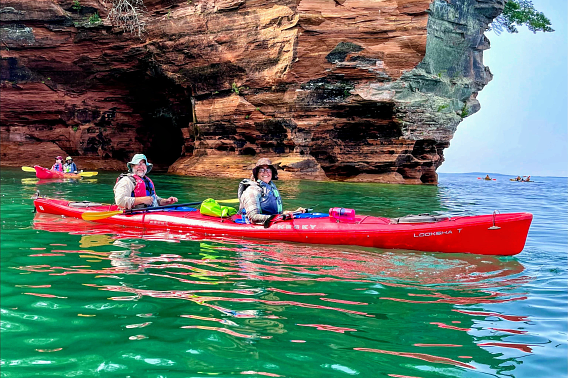Two smiling people in a tandem sea kayak pause in calm water in front of a tall sandstone sea cave. Behind them, another duo in a tandem sea kayak pause near the sea cave. Another set of kayakers are paddling around the headland.