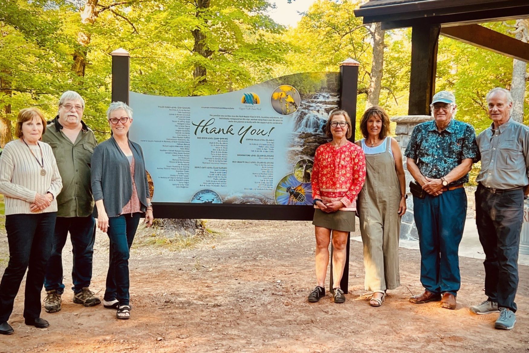 Seven people stand and flank a dedication sign outdoors.