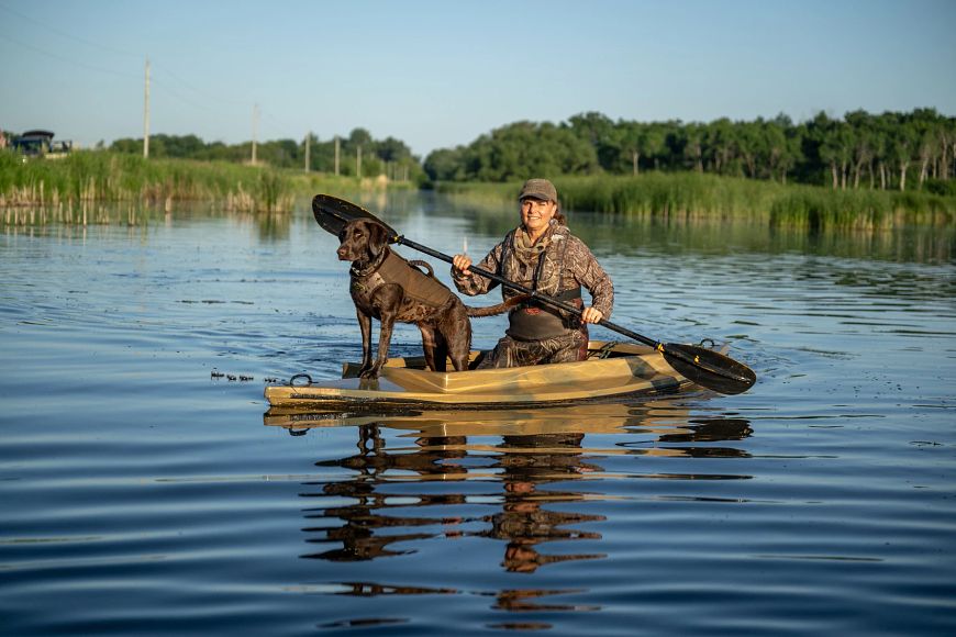 A women in camo paddles a duck skiff with a chocolate lab standing at attention on the prow.