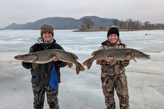 Two fisherwomen dressed for cold weather stand on the frozen water, smiling widely as they carefully hold the Northern Pike that each of them caught ice fishing. The two fish are nearly identical, with the one on the left just a bit bigger than the one on the right. The fishes’ heads are long with prominent underbites to their teeth-filled mouths. Their bodies are olive green with yellow undertones and distinct oblong light tan spots covering their sides. They have bold striping on their fins. In the background, bluffs rise steeply above the far bank of the frozen Mississippi River.