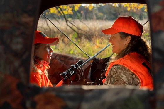 A woman and a child smile at each other while sitting in a deer blind.