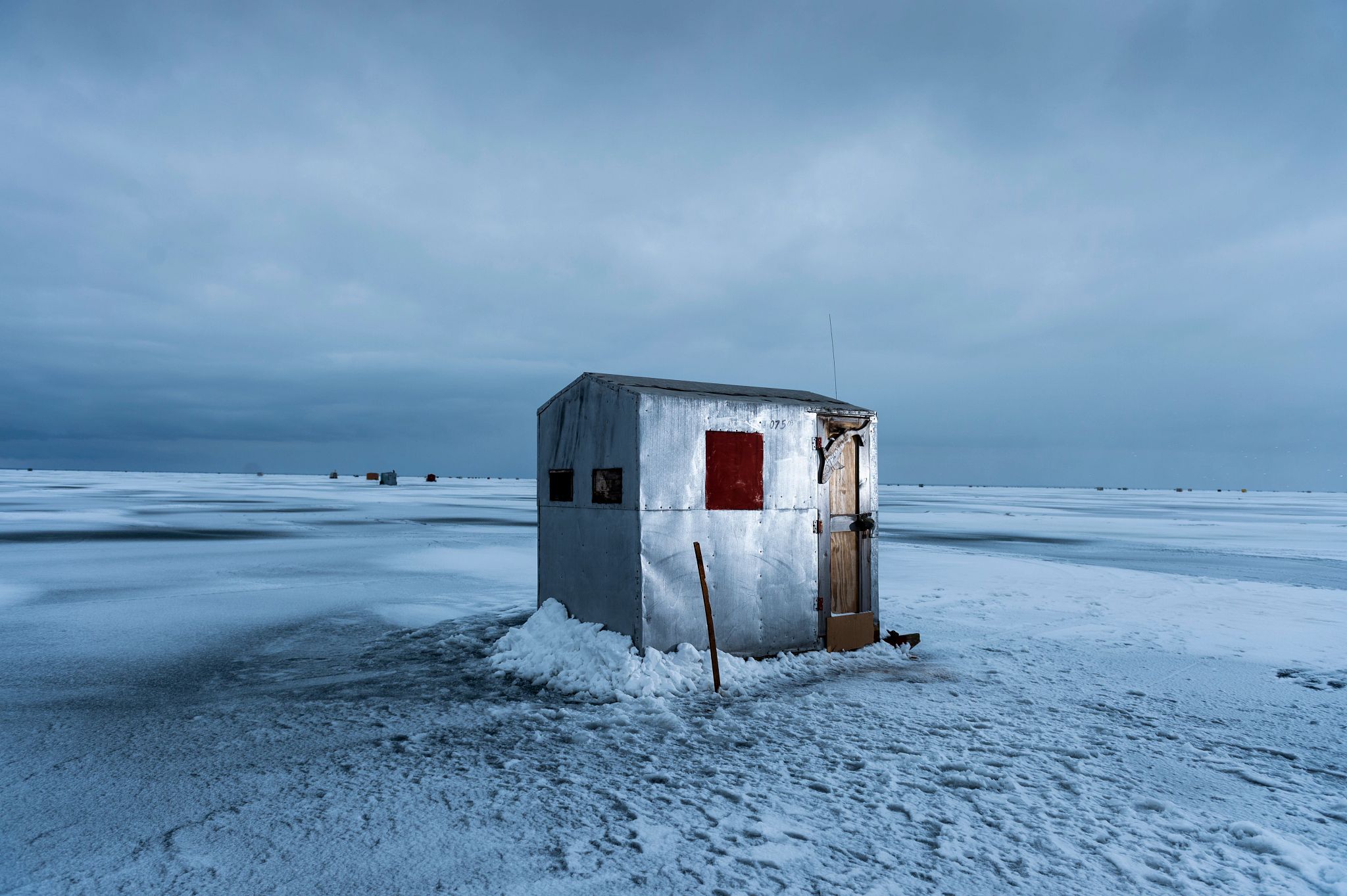 A silver ice shanty sits in the middle of an icy, snow-covered lake.