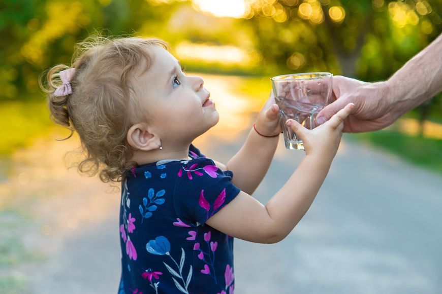 A young girl standing outside on a bright day grabs a glass of clear water from a hand out of frame.