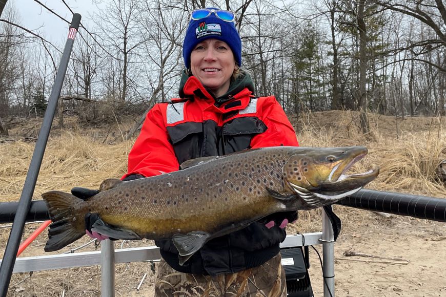 A women in a winter hat and coat holds a large brown trout in her hands and smiles.