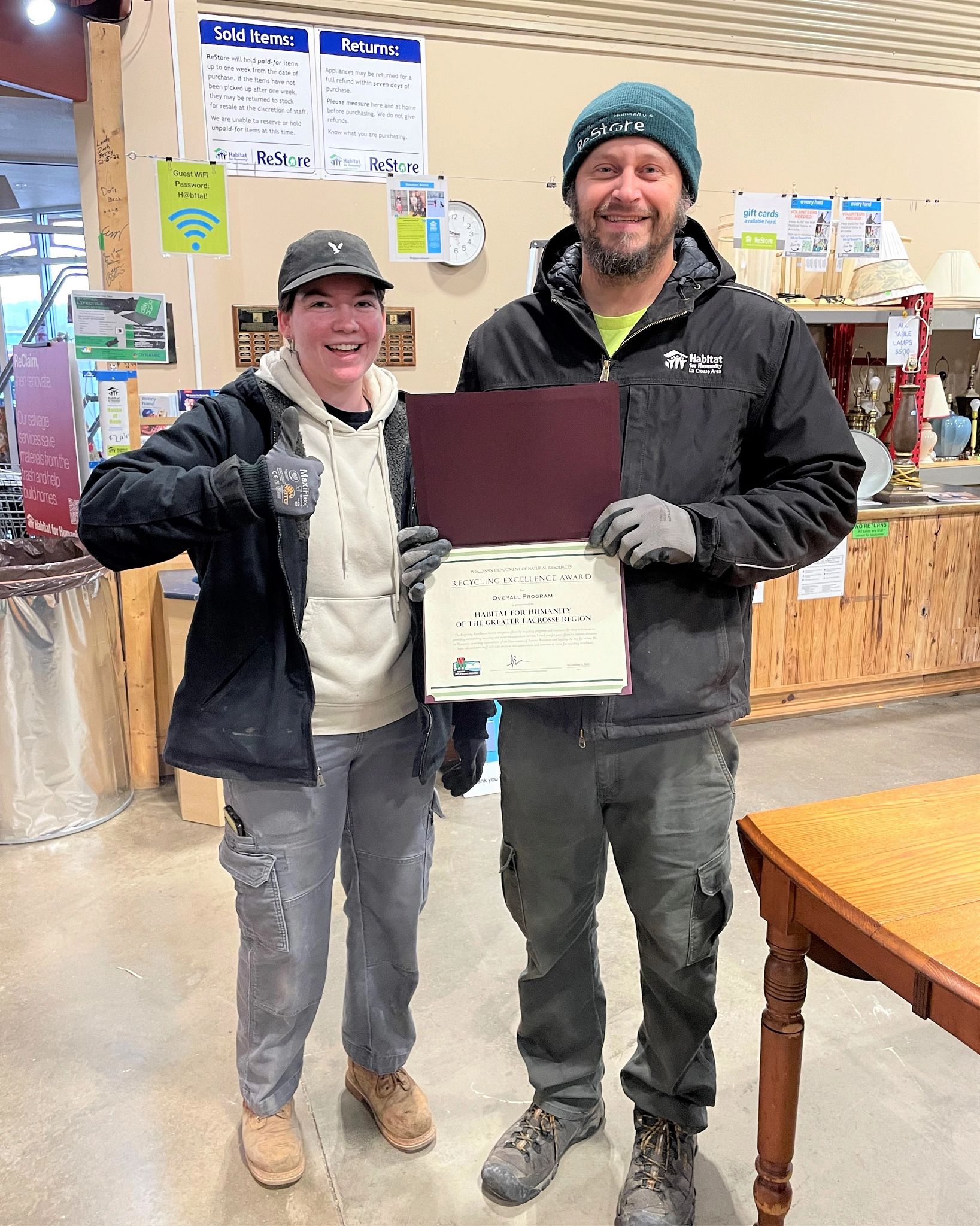 Two team members from Habitat for Humanity of the Greater La Crosse Region pose in a Habitat ReStore location with their 2023 Wisconsin Recycling Excellence Award certificate from the DNR. One team member offers a 'thumbs up' sign.