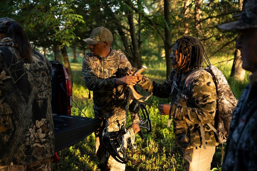 someone handing another person hunting tools from the back of a truck and other people standing nearby wearing camoflauge