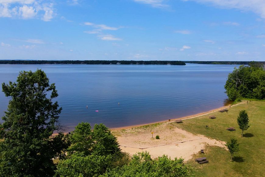 An aerial view of a large, bright blue lake with a grassy shoreline and beach in the foreground.