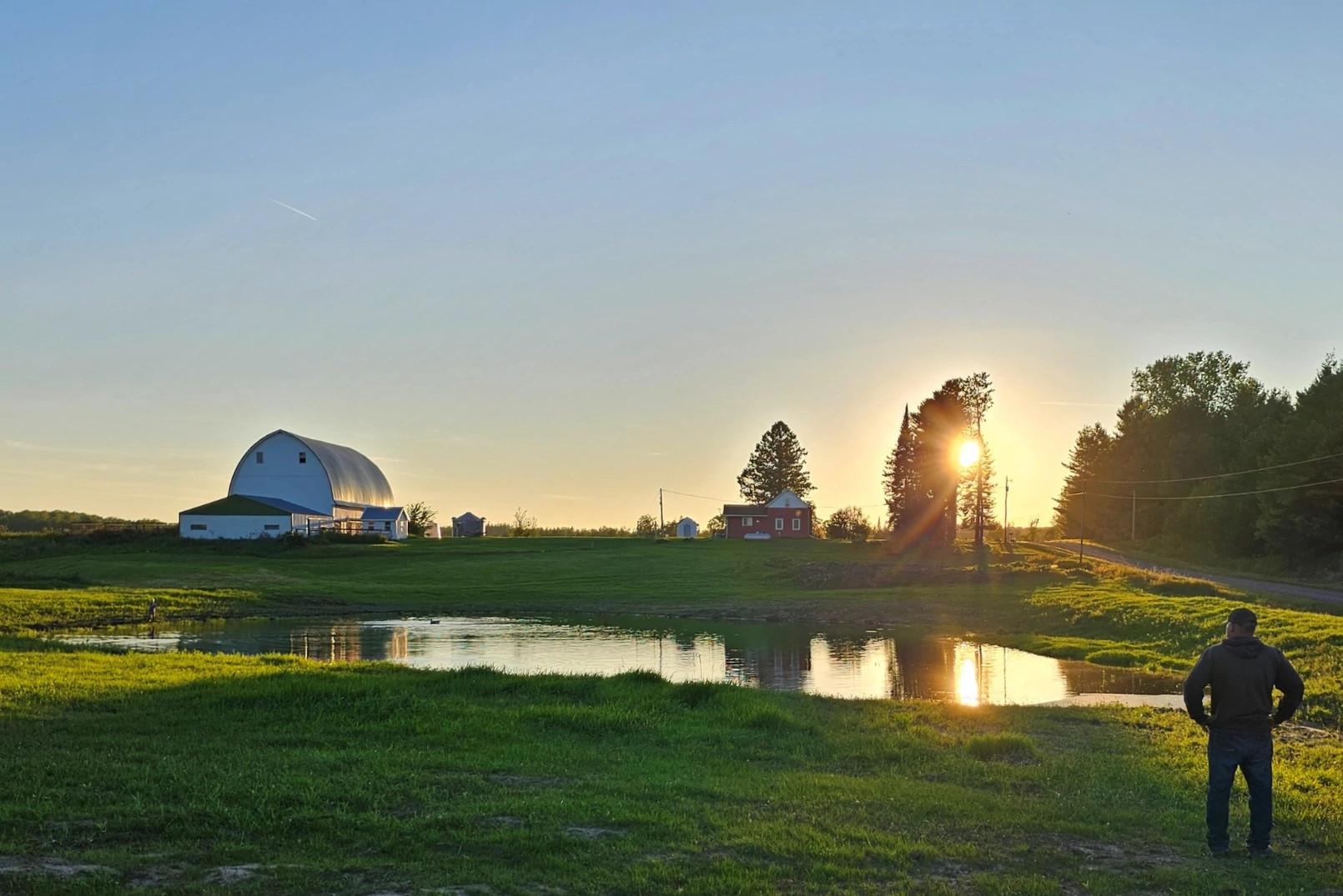 A man stands in front of a pond and surveys his farm, as the sun rises.. 