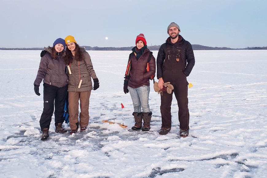 A group of individuals pose for a picture around a tip up while ice fishing. 