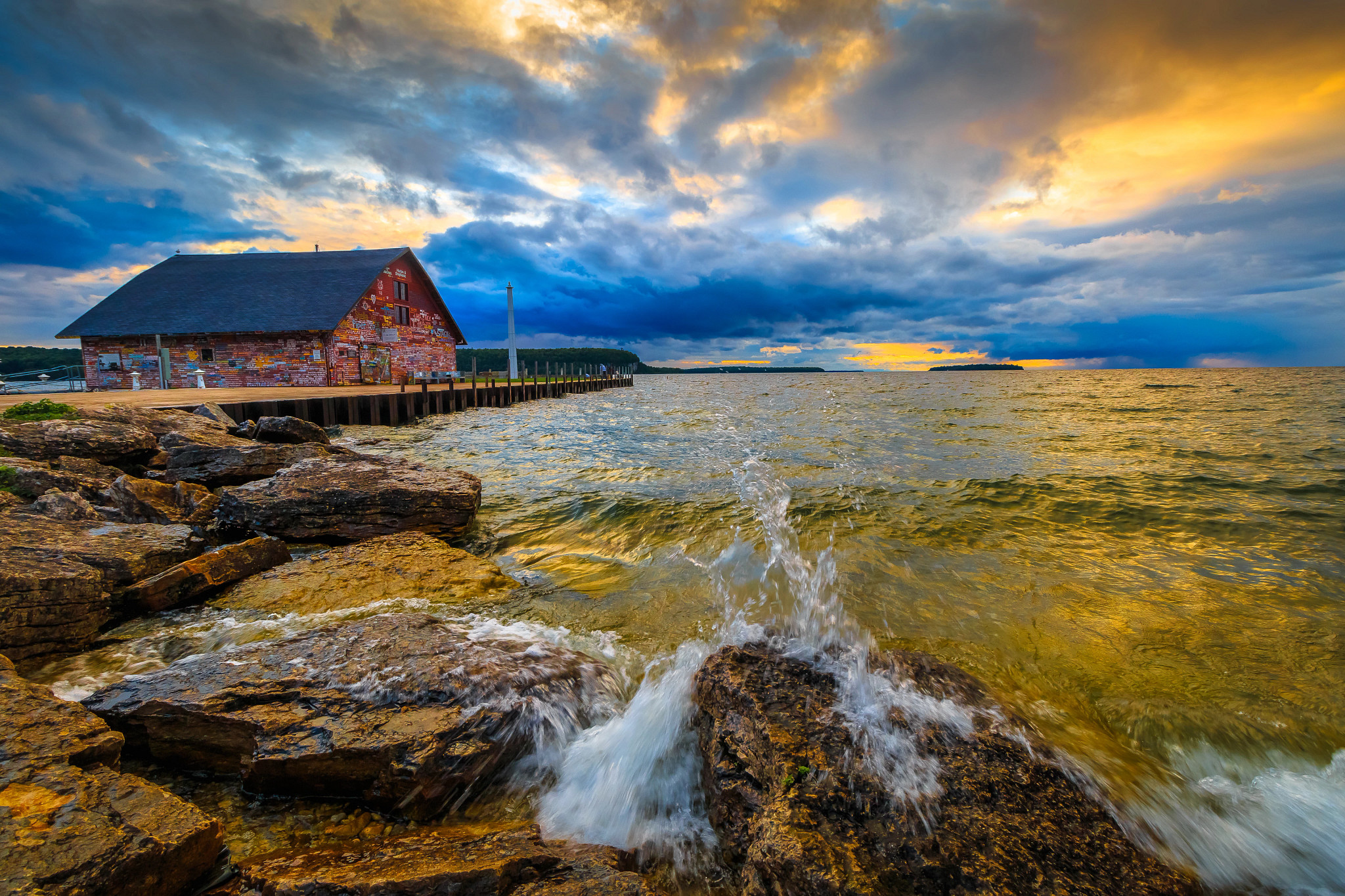 Golden rays of the sun attempt to burst through deep blue roiling clouds, heavy with rain. The waters reflect a strange brew of colors, tinted greenish yellow, as they crash on the rocky shore of Eagle Harbor. The Anderson Dock warehouse building is to the left of the approaching storm cloud. The red clapboard siding, strewn with colorful graffiti, makes this historical landmark stand out at the end of a long pier. 
