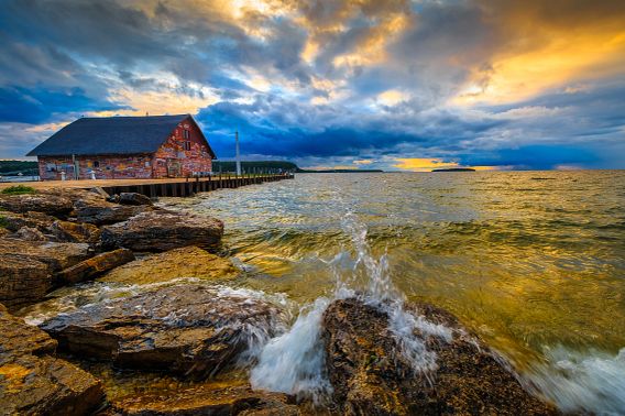 Golden rays of the sun attempt to burst through deep blue roiling clouds, heavy with rain. The waters reflect a strange brew of colors, tinted greenish yellow, as they crash on the rocky shore of Eagle Harbor. The Anderson Dock warehouse building is to the left of the approaching storm cloud. The red clapboard siding, strewn with colorful graffiti, makes this historical landmark stand out at the end of a long pier.