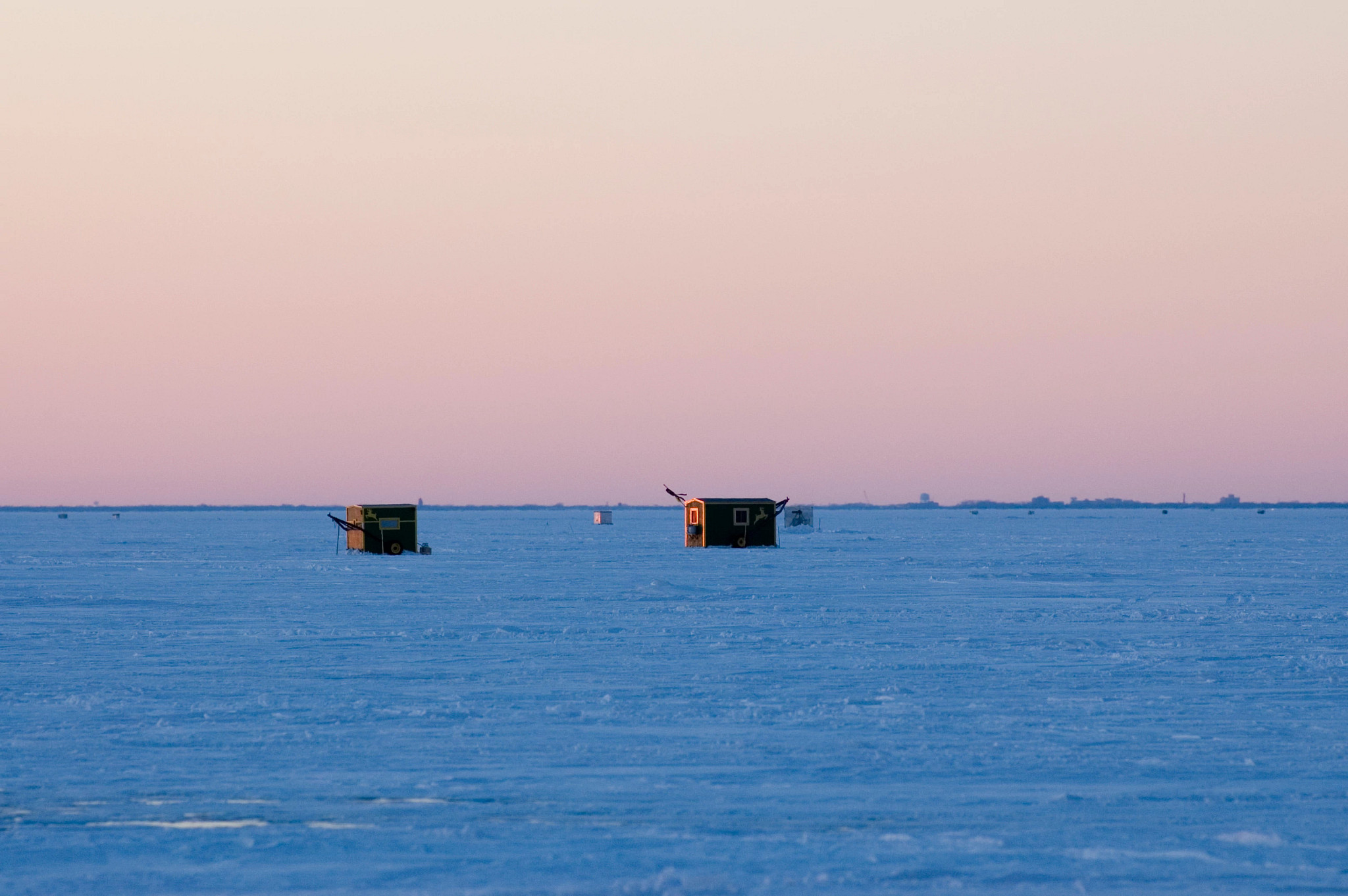 Two ice shanties sitting on the ice around sunrise or sunset.